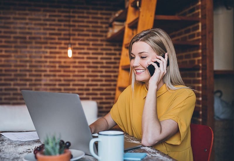 Blonde woman sitting at table in her modern loft apartment talking on a cellphone and using a laptop